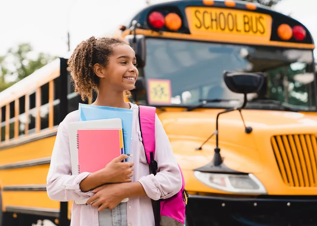 Girl holding notebooks standing next to a school bus