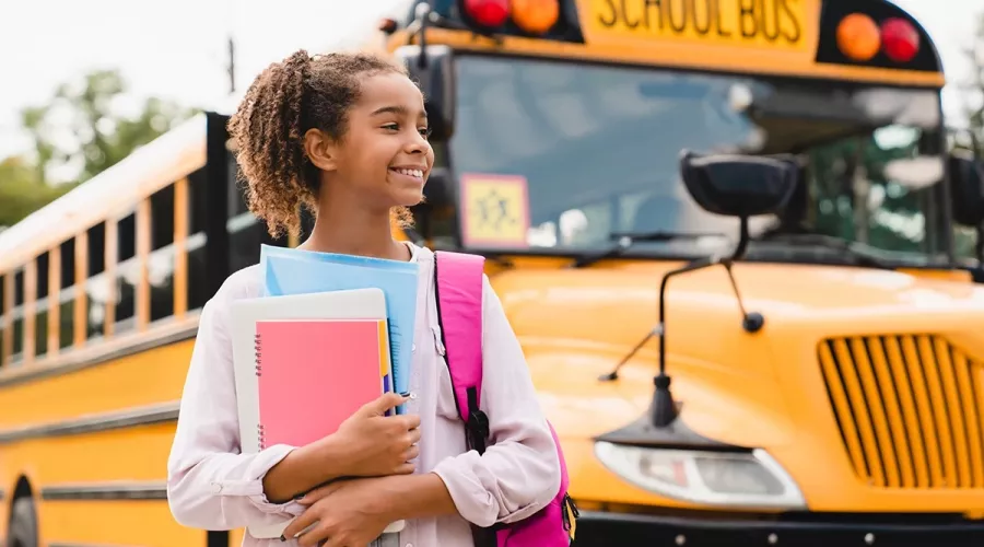 Girl holding notebooks standing next to a school bus